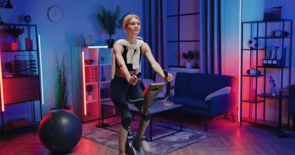 Mooie actieve sportieve slanke blonde in sportkleding oefening op de fiets trainer tijdens de avond thuis workout, gezonde levensstijl concept — Stockvideo