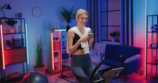 Милая улыбающаяся довольная активная спортивная стройная молодая женщина в спортивной одежде читает новости на смартфоне во время занятий на стационарном велосипеде дома вечером — стоковое видео