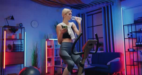 Активная и здоровая концепция образа жизни, где довольно спортивная стройная молодая женщина в спортивной одежде упражняется на стационарном велосипеде и питьевой воде во время домашней тренировки — стоковое видео