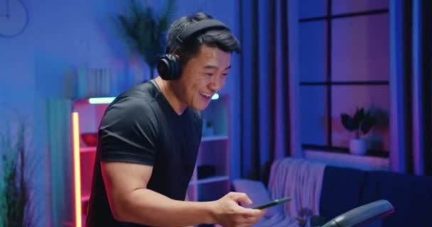 Aantrekkelijke vrolijke actieve atletische aziatische man in sportkleding luisteren moderne muziek in hoofdtelefoon tijdens het sporten op loopband tijdens de avond thuis workout, slow motion — Stockvideo