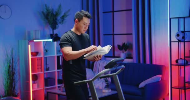Çekici, kendine güvenen, spor kıyafetli modern Asyalı adam koşu bandında yürüyor ve akşam evde çalışırken ilginç kitaplar okuyor, ağır çekimde. — Stok video