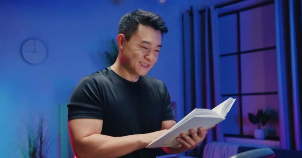 Gros plan de sympathique sportif positif homme asiatique de 30 ans en vêtements de sport qui lecture livre intéressant lors de l'exercice sur piste de course à la maison le soir — Video