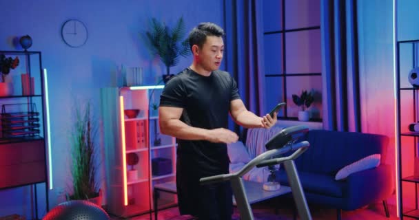 Beau sourire actif sportif asiatique guy dans sportswear marche sur tapis roulant à la maison dans la soirée et parler avec ami via appel vidéo sur smartphone — Video