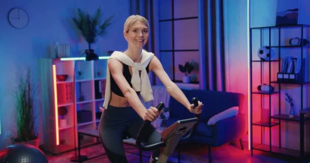 Портрет очаровательной улыбающейся активной спортивной девушки в спортивной одежде, которая поворачивает педали на стационарном велосипеде и показывает большие пальцы вверх в камеру в комнате с ночным освещением — стоковое видео