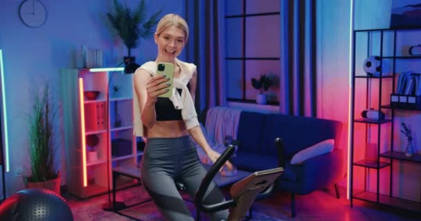 Досить усміхнена спортивна струнка молода жінка в спортивному одязі вправляється на стаціонарному велосипеді і робить смішне селфі на мобільному телефоні під час вечірнього домашнього тренування — стокове відео