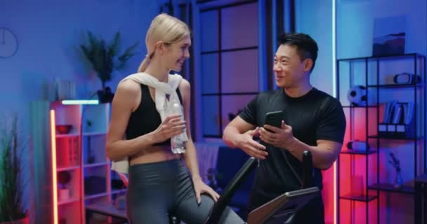 Schöner lächelnder glücklicher asiatischer Kerl in Sportbekleidung im Gespräch mit seiner hübsch fitten athletischen blonden Freundin, die abends auf dem Trainer-Fahrrad im schön dekorierten Raum trainiert — Stockvideo