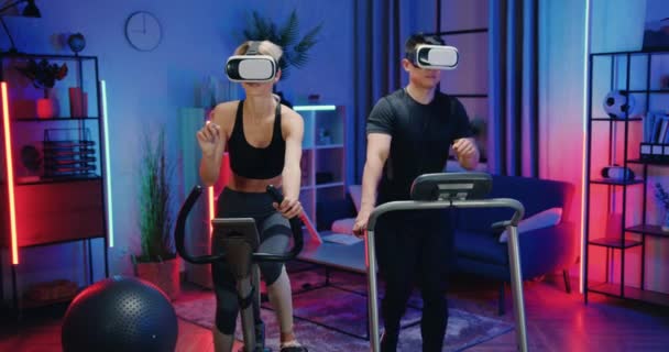 Attraktives, konzentriertes, sportlich-modernes multiethnisches Paar, das abends mit Augmented-Reality-Brille zu Hause auf Laufbahn und Fahrradtrainer trainiert — Stockvideo