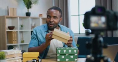 Çekici gülümseyen esrarengiz sakallı Afrikalı Amerikalı blogcu ev içi arka planda internet izleyicileri için video kaydederken kamerada karton kutular gösteriyor