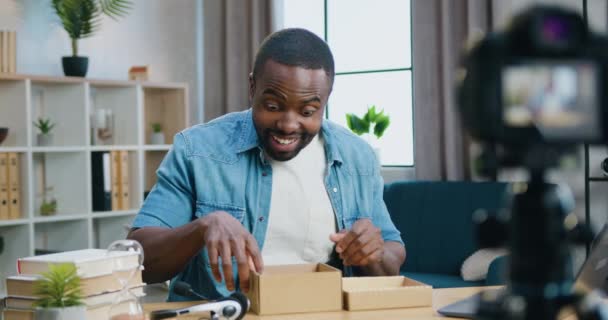 Bello felice sorridente blogger afro-americano barbuto facendo grandi occhi di sorpresa quando apri la scatola e tiri fuori un nuovo telefono durante lo streaming sulla fotocamera da casa per gli spettatori online — Video Stock