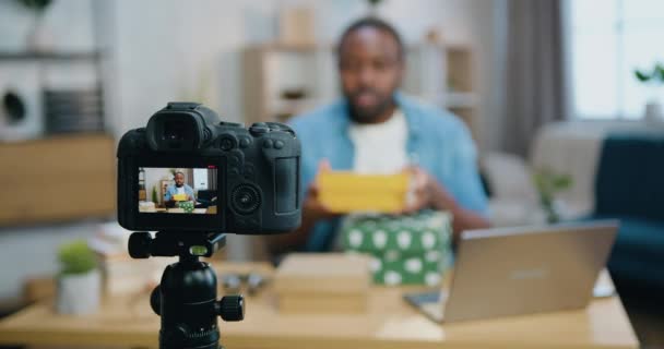 Вид через камеру на красивый положительный загадочный бородатый африканский парень, который записывает новое видео для своего видеоблога, показывая коробки с неизвестными подарками — стоковое видео
