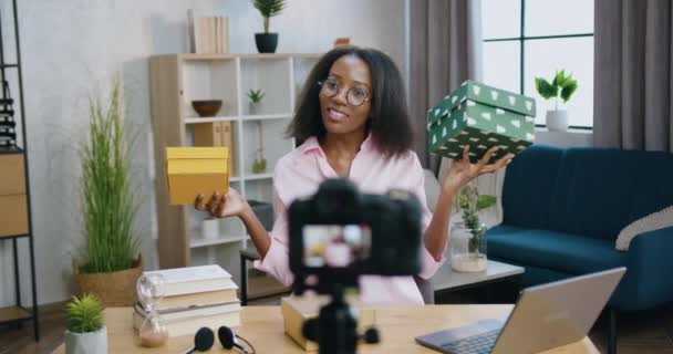 Довольно улыбающаяся, довольная молодая африканская блоггерша держит в руках подарочные коробки во время записи нового видеоблога для интернет-аудитории в домашней студии — стоковое видео
