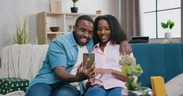 Bonito sorriso feliz amoroso jovem casal afro-americano sentado no sofá em casa e falando no smartphone durante o bate-papo por vídeo com amigos ou parentes — Vídeo de Stock