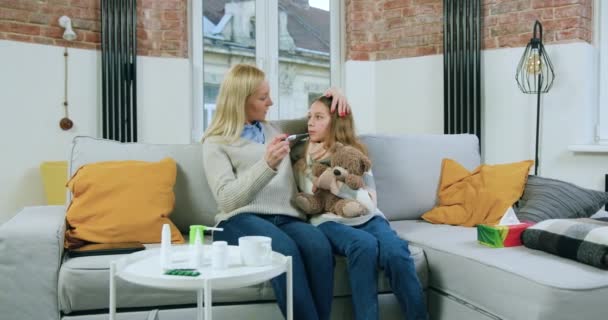 Atractiva madre positiva segura de 40 años midiendo la temperatura de su hija adolescente que está sentada cerca de ella en un sofá suave, concepto de gripe — Vídeo de stock