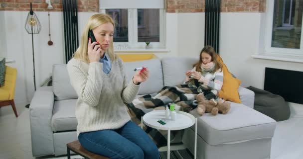 Schattig zielig ziek tiener meisje met sjaal rond hals liggend op comfortabele bank thuis, hoesten en het drinken van warme thee, terwijl haar bezorgd moeder heeft mobiele gesprek met arts — Stockvideo