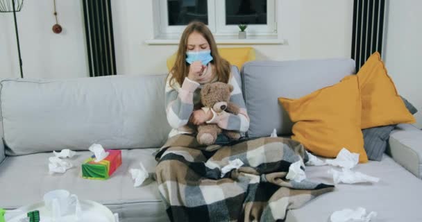 Приятная грустная измученная девочка-подросток в маске с шарфом на шее, сидящая на мягком диване и кашляющая, потому что простудилась и получала лечение дома, закрыть — стоковое видео