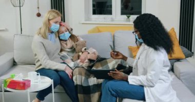 Sevimli, siyah tenli, maskeli genç bir kadın doktor, koruyucu maskeli, koruyucu maskeli 15 yaşındaki annesine ve hasta kızına tablet veriyor.