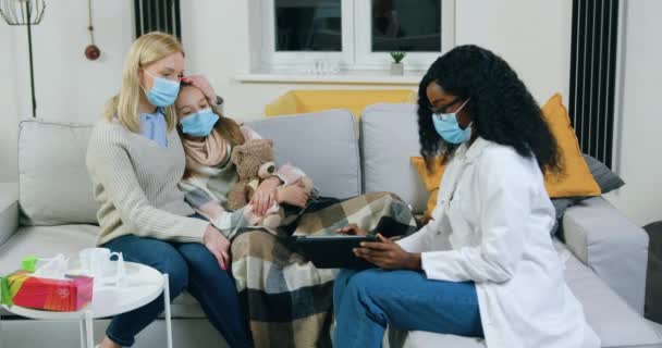 Aangename ervaren Afrikaanse Amerikaanse vrouwelijke arts in beschermende masker bezoeken zieke tiener patiënt die onder toezicht van moeders thuis en het geven van instructies over verdere methoden van behandeling — Stockvideo