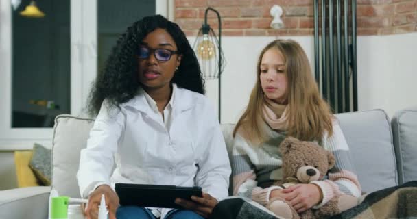 Nahaufnahme der schönen selbstbewussten, qualifizierten schwarzhäutigen Hausärztin mit Brille, die ihre kranke Teenie-Patientin zu Hause besucht und Anweisungen gibt, wie sie Tabletten einnimmt — Stockvideo