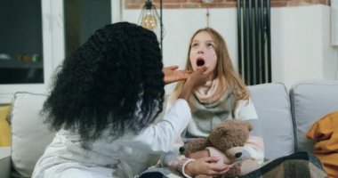 Sevimli deneyimli siyah tenli kadın özel doktor ev ziyareti, hastalık ve sağlık sigortası konsepti sırasında genç kızların boğazını kontrol ediyor.