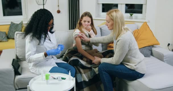 Atraktivní sebevědomá žena držící se za ruku své vyděšené dospívající dívky, která zkušená africká americká lékařka by měla podat injekci během domácí návštěvy, nemoci a zdravotní péče — Stock video