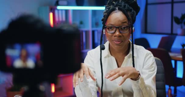迷人而自信的有经验的非洲裔美国女人，在晚上的办公室背景下，带着无线耳机在摄像机上录音 — 图库视频影像