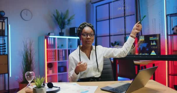 Påskyndad fotografering av sympatisk positiv säker skicklig afrikansk amerikansk kvinna i glasögon som förklarar export och import indikatorer ritade på glasskiva på kameran på kontoret vid sen tidpunkt — Stockvideo