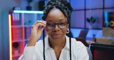 Akşam çalışma odasının arka planında gözlüğünü çıkarırken kameraya bakan ve gülümseyen Afro-Amerikan kadın ofis müdürünü yakından çek.