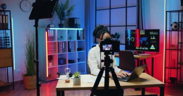 Conceito de escritório Podcast onde atraente positiva confiante mulher de negócios africana habilidosa em óculos e desgaste elegante trabalhando no laptop no escritório de tempo escuro simultaneamente gravação de vídeo — Vídeo de Stock