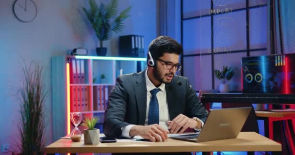 Trolig säker erfaren smart skäggig affärsman i kostym bär headset sitter framför laptop under videomöte på kvällen arbetsrum bakgrund — Stockvideo