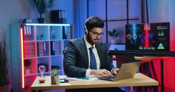 Zakelijk concept waar sympathieke ernstige zelfverzekerde ervaren bebaarde office manager met headset verblijf in de avond om zijn computerwerk af te maken — Stockvideo