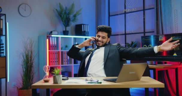 Привлекательный улыбающийся удовлетворенный успешный современный бородатый бизнесмен в костюме имеет приятный телефонный разговор во время работы в вечернем офисе, замедленная съемка — стоковое видео