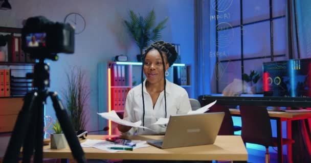 Krásná spokojená úspěšná africká americká žena se stylovým účesem drží online chat se svým internetovým publikem a zvrací listy papírů na konci proudu — Stock video
