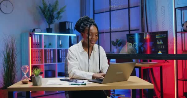Очень позитивная уверенная в себе целеустремленная опытная африканская бизнесвумен, сидящая на своем рабочем месте в вечернем офисе и сидящая за компьютерной клавиатурой, концепция рабочего процесса — стоковое видео