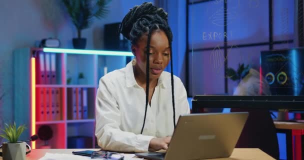 Hermosa mujer de negocios afroamericana responsable y segura de sí misma con un peinado elegante que trabaja en la computadora en su lugar de trabajo en la oficina de la noche, de cerca, en cámara lenta — Vídeos de Stock