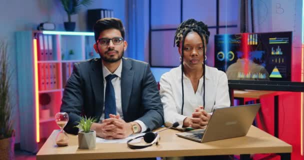 Attrayant positif amical homme d'affaires barbu moderne hautement qualifié et sa collègue afro-américaine posant à la caméra avec des sourires sincères dans le bureau du soir — Video