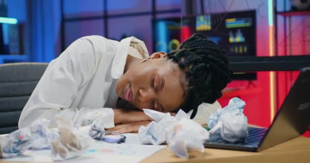 美しい疲れて過労のアフリカ系アメリカ人女性事務員が職場で寝ている夜のオフィスの背景に多くの崩壊した論文を置く — ストック動画
