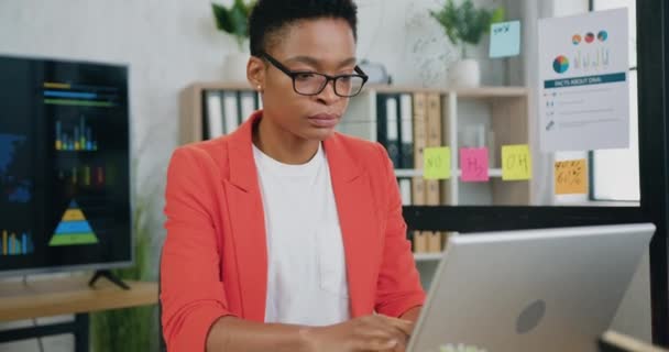 Mujer africana joven enfocada que trabaja en el ordenador portátil mientras está sentada en el lugar de trabajo — Vídeos de Stock