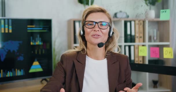 Portret close-up van een glimlachende jonge blanke vrouwelijke callcenter werknemer met telefoon headset en iets praten op online camera, in modern kantoor — Stockvideo