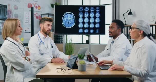 Привлекательные квалифицированные многонациональные врачи мужского и женского пола, сидящие за столом заседаний в медицинском кабинете и обсуждающие пациентов с МРТ мозга, медицинскую концепцию — стоковое видео
