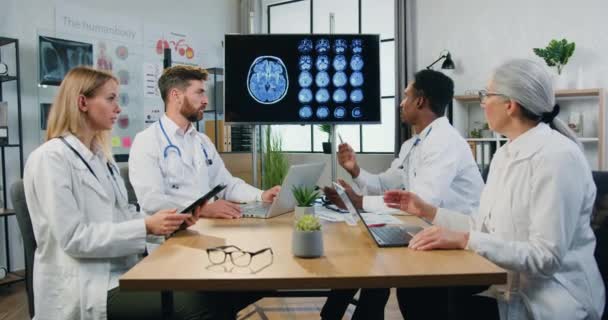 Симпатичная, уверенная в себе, опытная мультикультурная команда неврологов проводит совместное заседание в медицинском кабинете по поводу лечения пациентов с использованием результатов томографии — стоковое видео