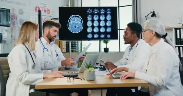 Dobře vypadající sebevědomí, pracovití multikulturní lékaři sedící kolem stolu v moderní klinice a seznamující se se zprávou, zpomalený pohyb — Stock video