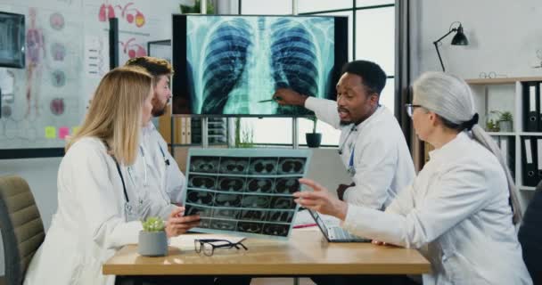 Attraente successo intenzionale esperto multietnico quattro medici che esaminano i pazienti radiografia del torace e polmoni scansione durante briefing congiunto in studio medico — Video Stock