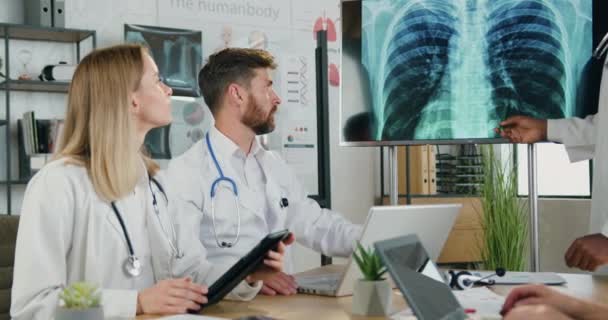 Goed uitziende ervaren mannelijke en vrouwelijke artsen die de resultaten van radiografie van de borst op digitaal scherm in het medisch kantoor onderzoeken tijdens een ontmoeting met andere collega 's — Stockvideo
