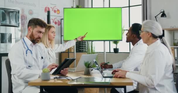 Os médicos multiétnicos bem sucedidos profissionais atraentes que prendem a reunião conjunta no escritório médico e usam o monitor digital com tela chave verde do croma — Vídeo de Stock