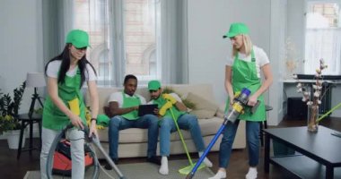 İyi görünümlü, çoklu etnik çeşitlilikte mutlu erkek-temizleyiciler tablet bilgisayarda oyun sonuçlarından keyif alırken, yetenekli kadın partnerleri müşterilerinde elektrikli süpürgeyle halı temizliyor.