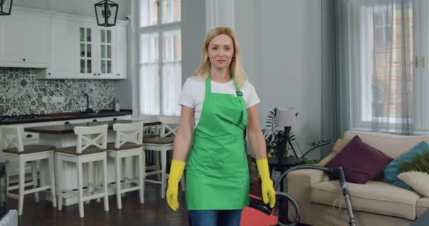 Όμορφη χαμογελαστή επαγγελματίας ενήλικας ξανθιά γυναίκα-εργαζόμενος της εταιρείας καθαρισμού στέκεται μπροστά από την κάμερα με σταυρωμένα χέρια στο σπίτι των πελατών — Αρχείο Βίντεο