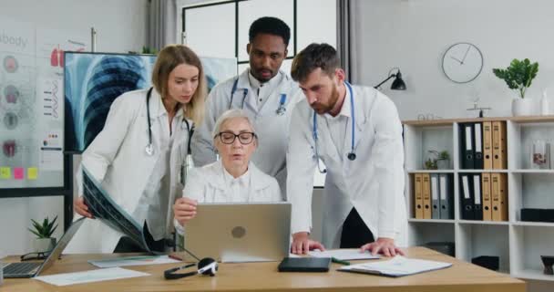 Medicii multietnici inteligenți și respectatul lor medic de succes care lucrează cu rezultate pe ecranul laptopului și scanarea cu raze X a plămânilor — Videoclip de stoc