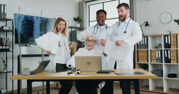 Atraktivní veselí spokojení úspěšní kvalifikovaní multietničtí doktoři, kteří dostávají dobré zprávy na obrazovce notebooku, rozhazují zprávy po hlavách, tleskají rukama a plácají si mezi sebou. — Stock video