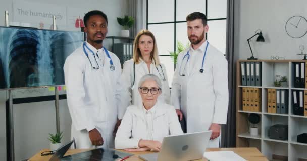 Atrakcyjny pozytywny sukces doświadczony wielonarodowy zespół lekarzy patrzących w kamerę w nowoczesnym gabinecie medycznym, widok z przodu — Wideo stockowe