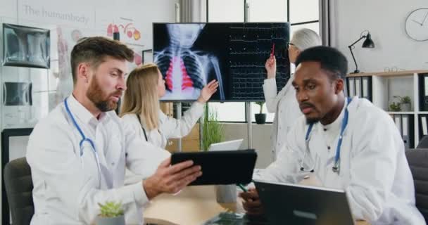 タブレットPCで2人の多民族男性医師が結果を修正し、 2人の女性医師がデジタル画面でハートビートの結果を議論する医療概念 — ストック動画